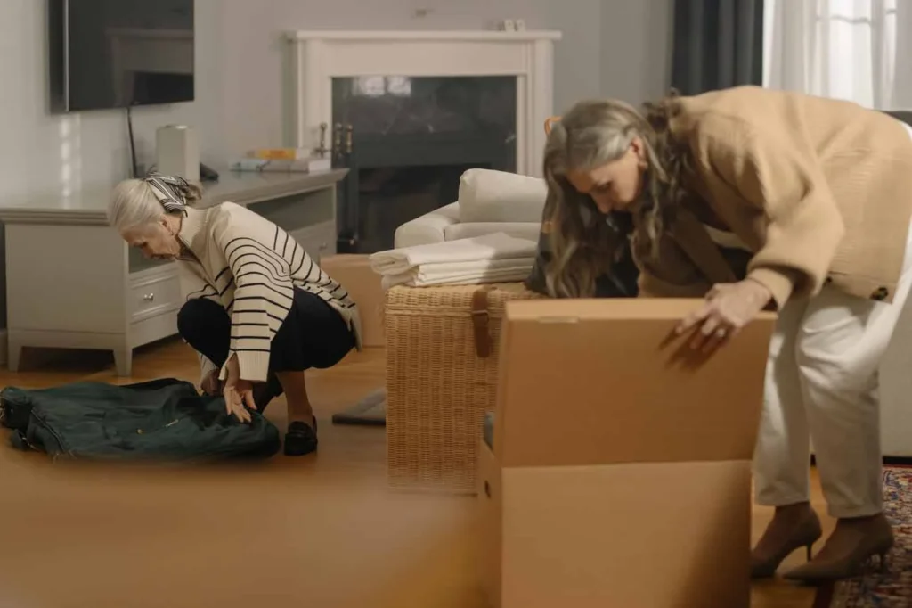 Two older women packing belongings into boxes in a living room before a move